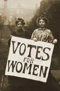 Old Black and white photo of two young women shading behind a large sign that reads "Votes for Women".
